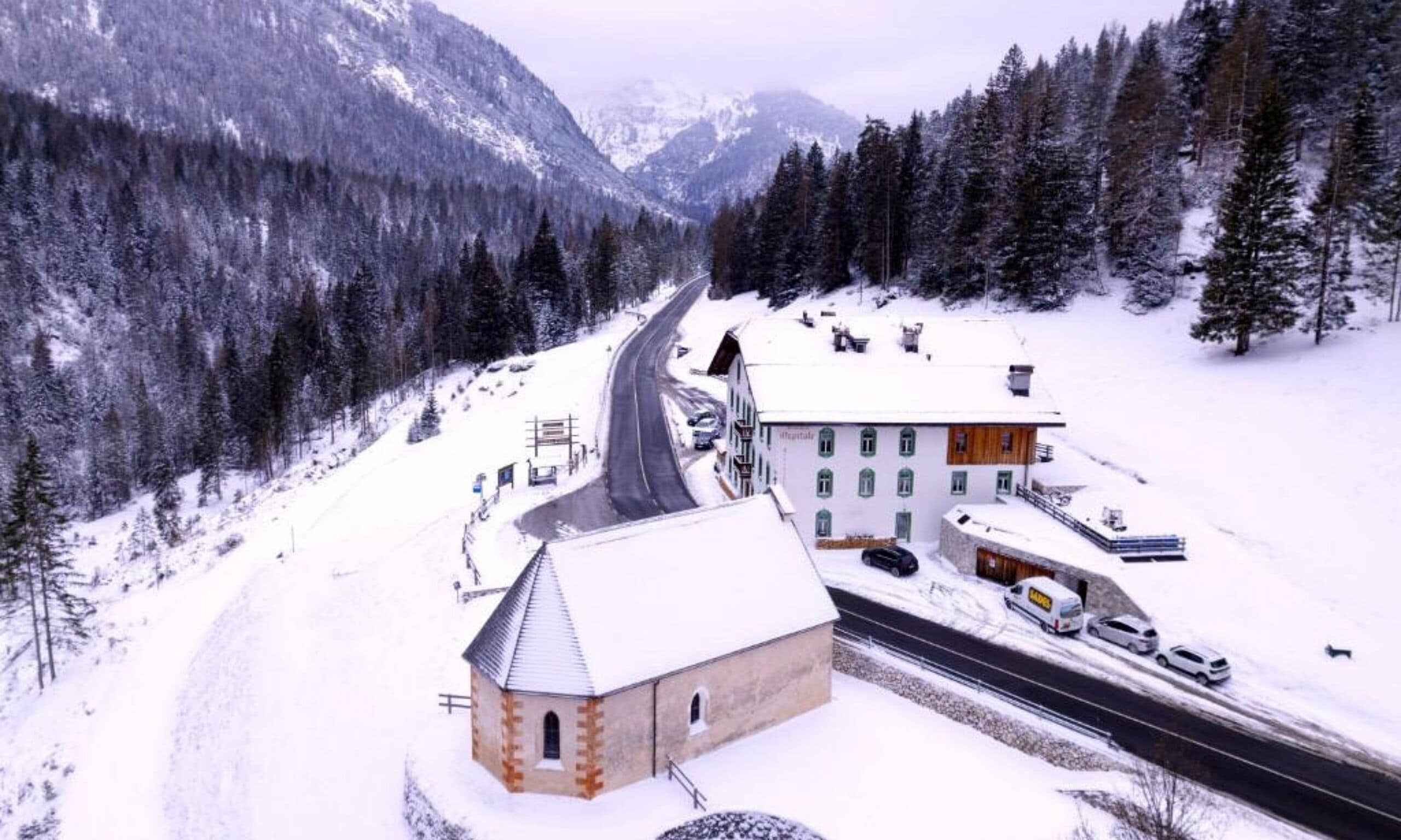 A snow-covered mountain village with a main road leading through it. There are buildings with snow on their roofs and a church-like structure with a bell tower.