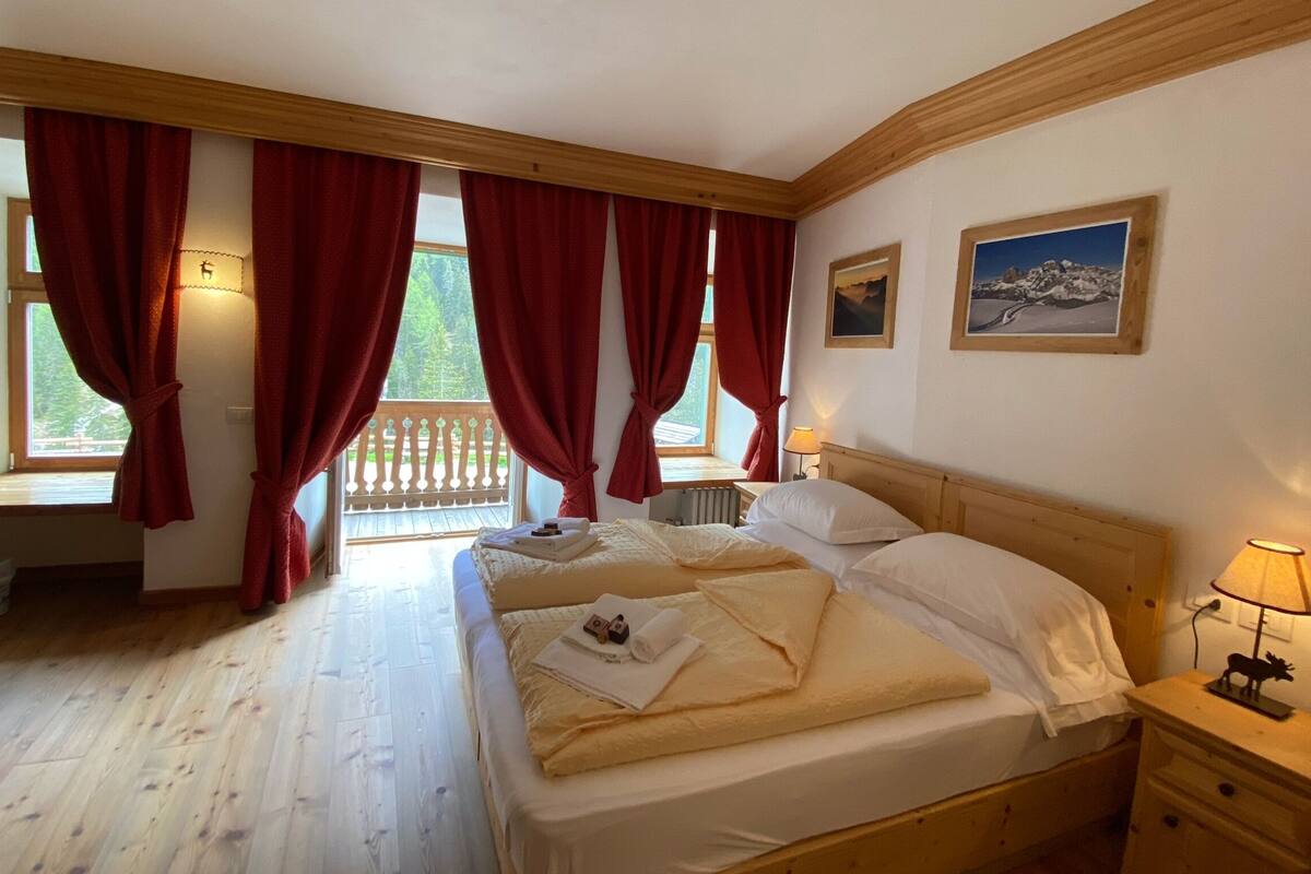 A bedroom with two beds, red curtains, a wooden headboard, a window with a view of greenery, and a small table with a lamp.