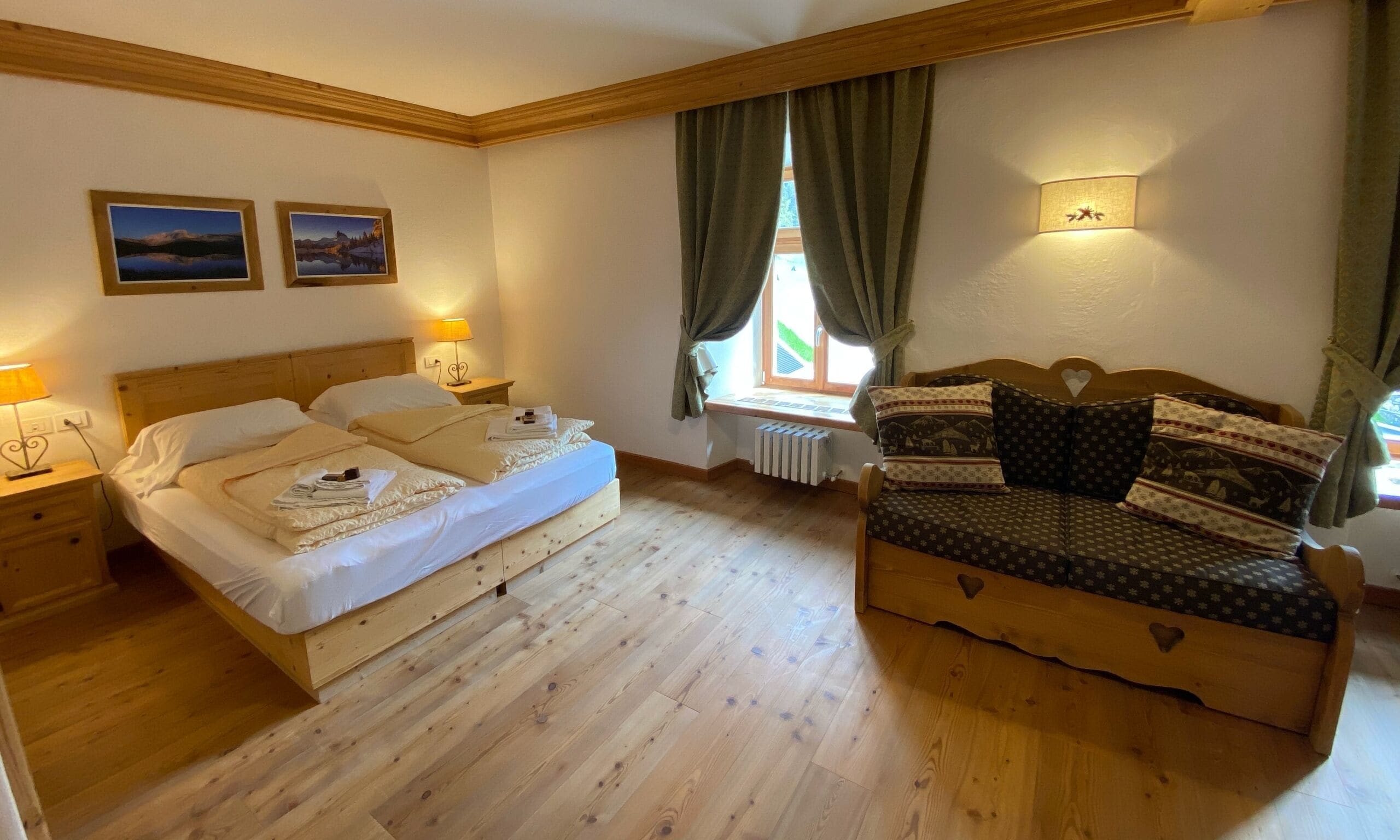 Un dormitorio con cama de madera, un sofá de madera, dos mesitas de noche, un ventanal con cortinas y dos cuadros colgados en la pared.