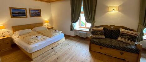 Un dormitorio con cama de madera, un sofá de madera, dos mesitas de noche, un ventanal con cortinas y dos cuadros colgados en la pared.
