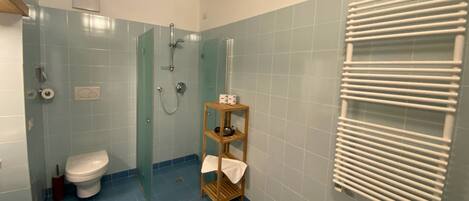A bathroom with a toilet, a glass shower enclosure, a wooden shelf with towels, and a white towel rack.