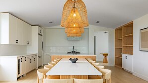 A modern kitchen with white cabinetry, a central island, and a dining area with a long table and chairs. - The Joyrise House at Solaire Lunara Bay (Key West)