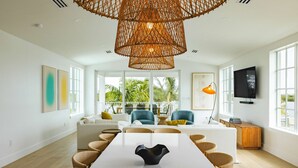 A spacious living area with a large dining table, comfortable seating, a wicker chandelier, and a view of palm trees through large windows. - The Joyrise House at Solaire Lunara Bay (Key West)