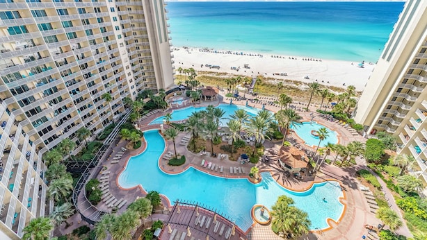 Un hotel de gran altura con un amplio área de piscina, que incluye una piscina central y una piscina circular más pequeña, rodeadas de palmeras y sillones de descanso.