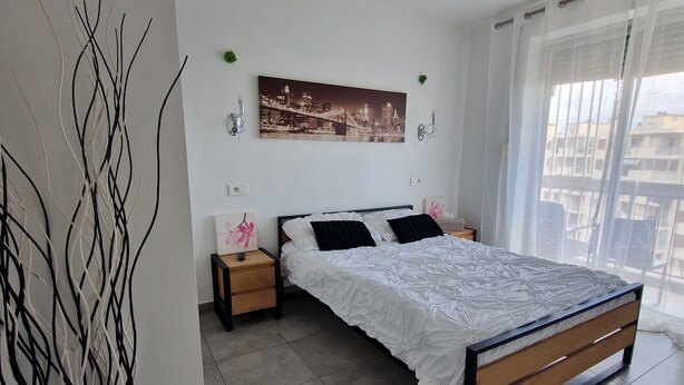 A bedroom with a large bed, a nightstand, a wall-mounted lamp, a cityscape painting, and a tall vase.