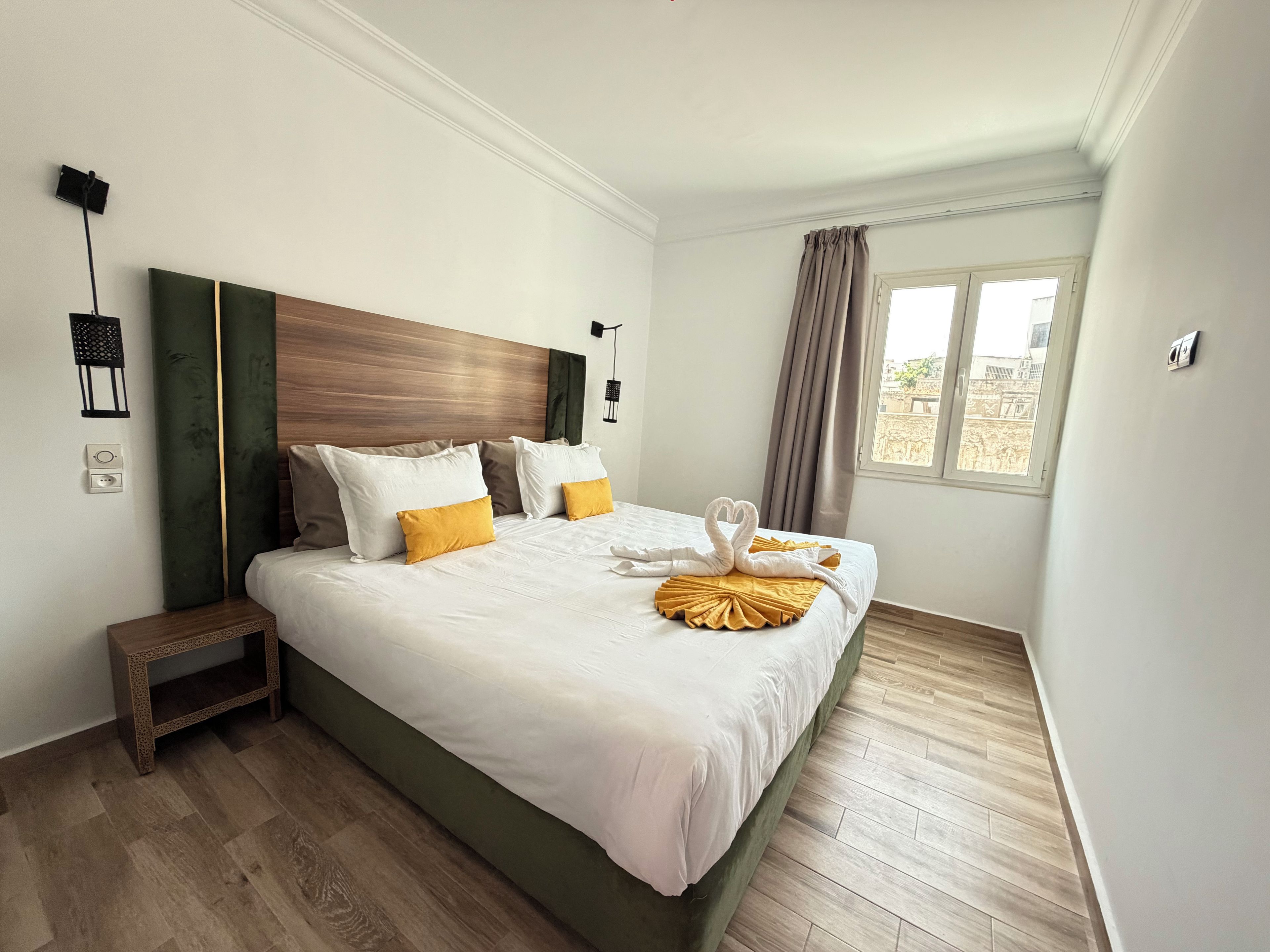 Habitación de hotel con una cama grande, cabecera de madera, mesita de noche, ventana con cortinas y vistas al exterior.