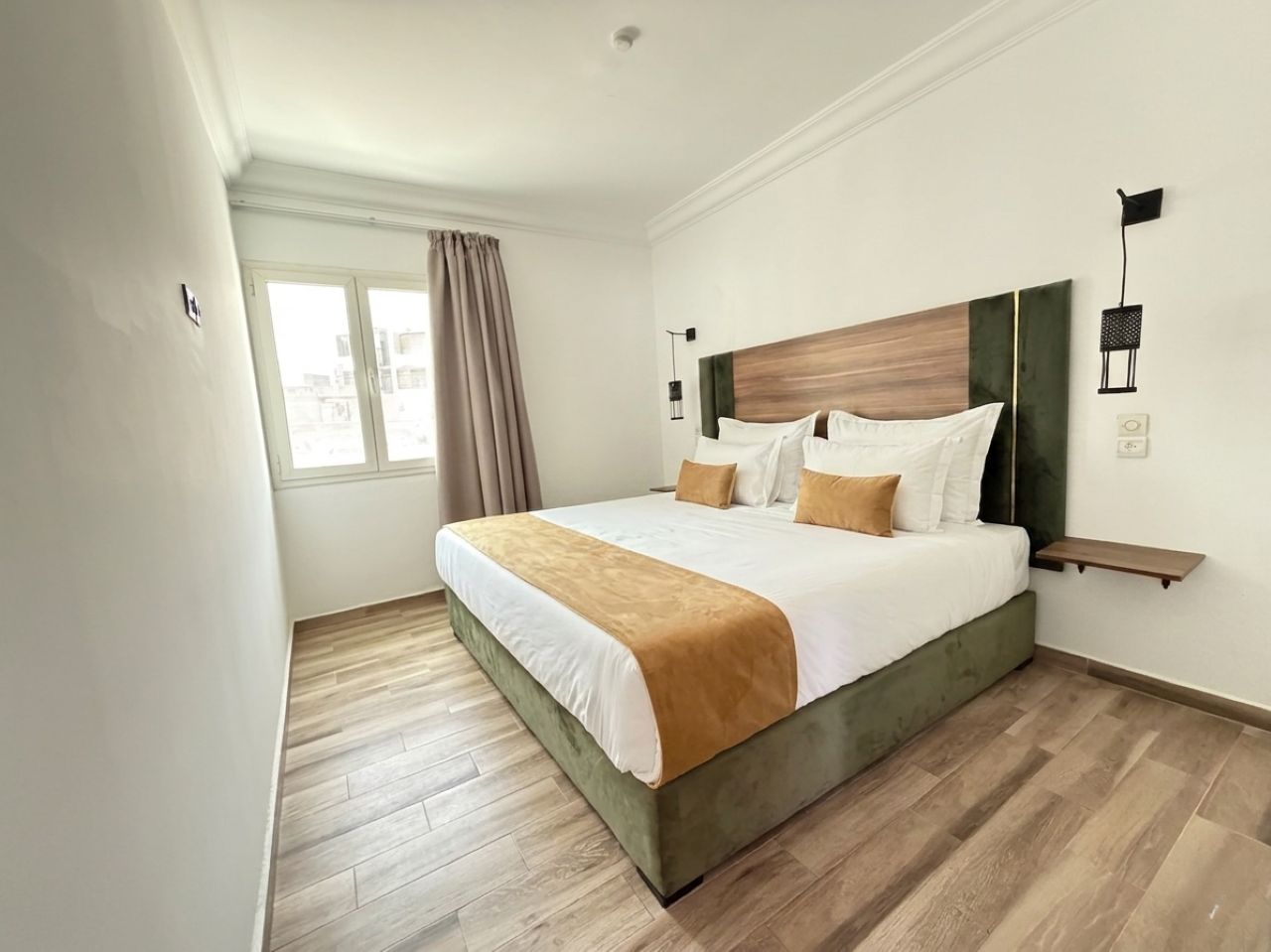 Une chambre d’hôtel avec un grand lit, une table de chevet, une fenêtre avec des rideaux et un luminaire fixé au mur.