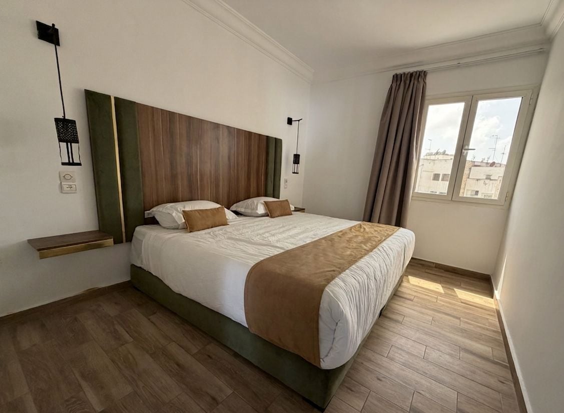 A hotel room with a large bed, wooden flooring, a headboard with a wooden panel, a window with curtains, and a wall-mounted light fixture.