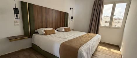 A hotel room with a large bed, wooden flooring, a headboard with a wooden panel, a window with curtains, and a wall-mounted light fixture.