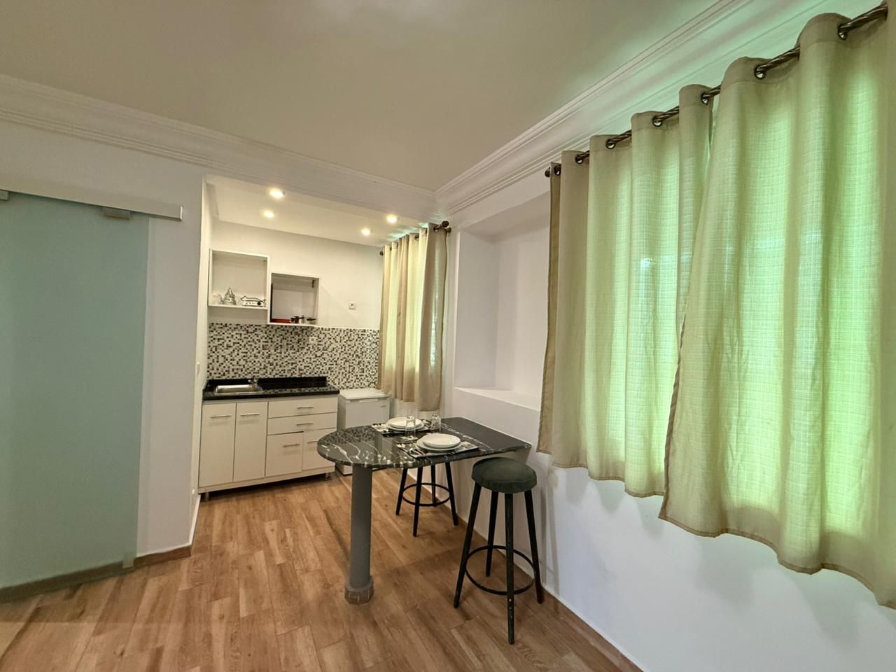 Una cocina compacta con comedor, que incluye una mesa, taburetes, un ventanal con cortinas y suelo de color claro.