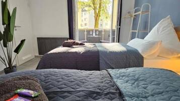 Ein Schlafzimmer mit zwei Betten, einem Fenster mit Blick auf Gebäude und einen Baum, einem Heizkörper und einer Pflanze.