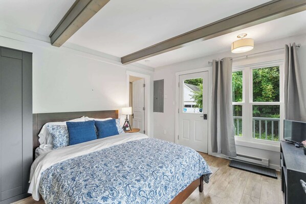 A bedroom with a bed, a desk, a window with curtains, and a door.