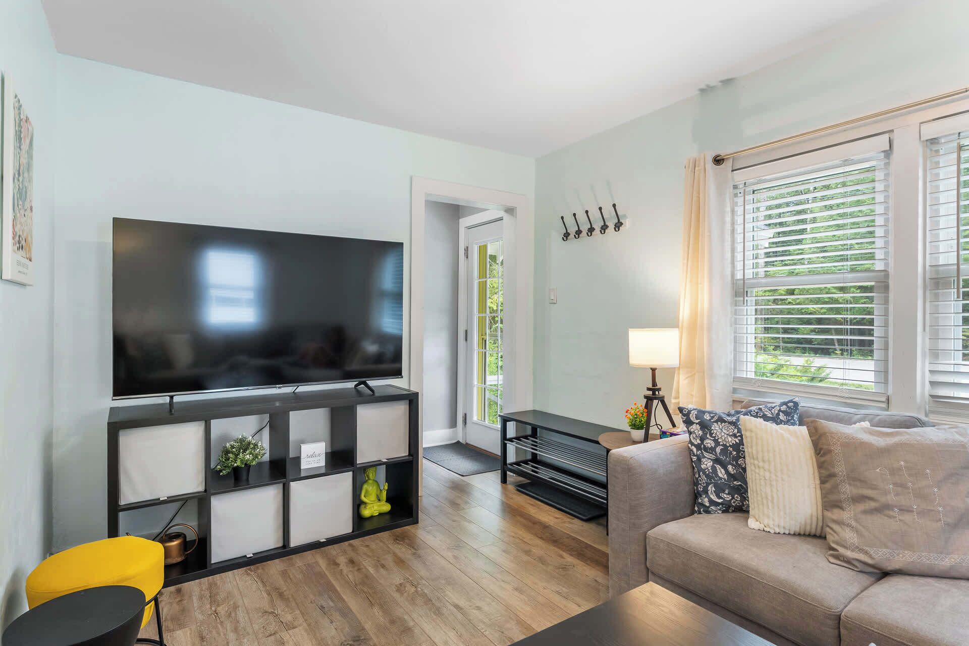 A living room with a flat-screen TV, a sofa, a yellow chair, a black console table, and a window with blinds.