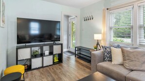 Ein Wohnzimmer mit Flachbildfernseher, einer Couch, einem gelben Sessel, einem schwarzen Konsolentisch und einem Fenster mit Jalousien.