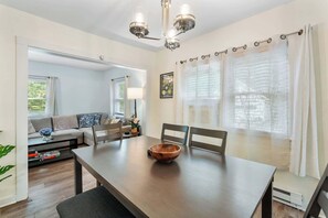 A dining area with a wooden table, chairs, and a bowl. There is a sofa and a window with curtains in the background.