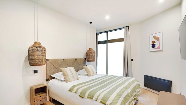Une chambre avec un grand lit, deux tables de chevet, une fenêtre avec des rideaux, un radiateur fixé au mur et un luminaire suspendu.