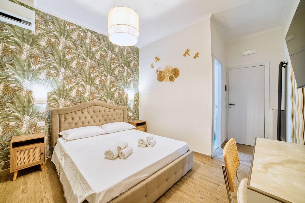 A bedroom with a large bed, wooden nightstands, a hanging light fixture, and a tropical wallpaper design.