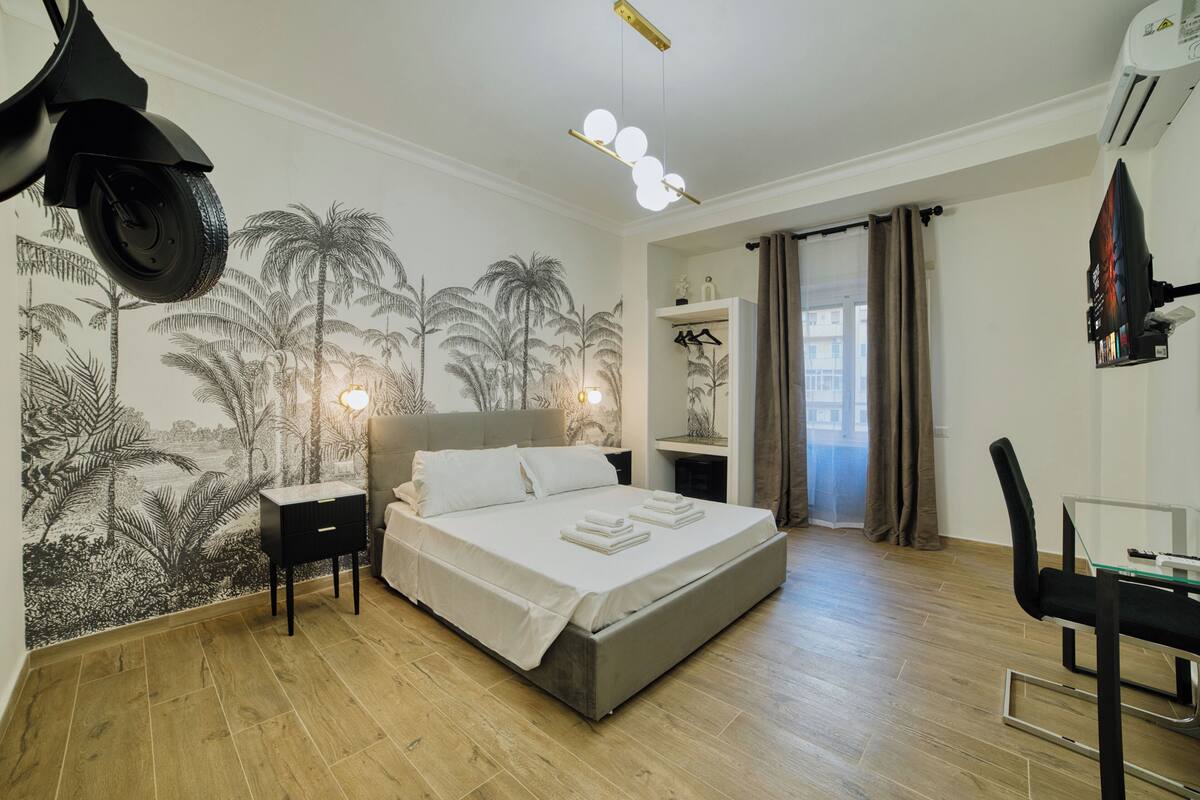 A bedroom with a bed, bedside tables, a desk, and a chair. There is a mural on the wall and a window with curtains.