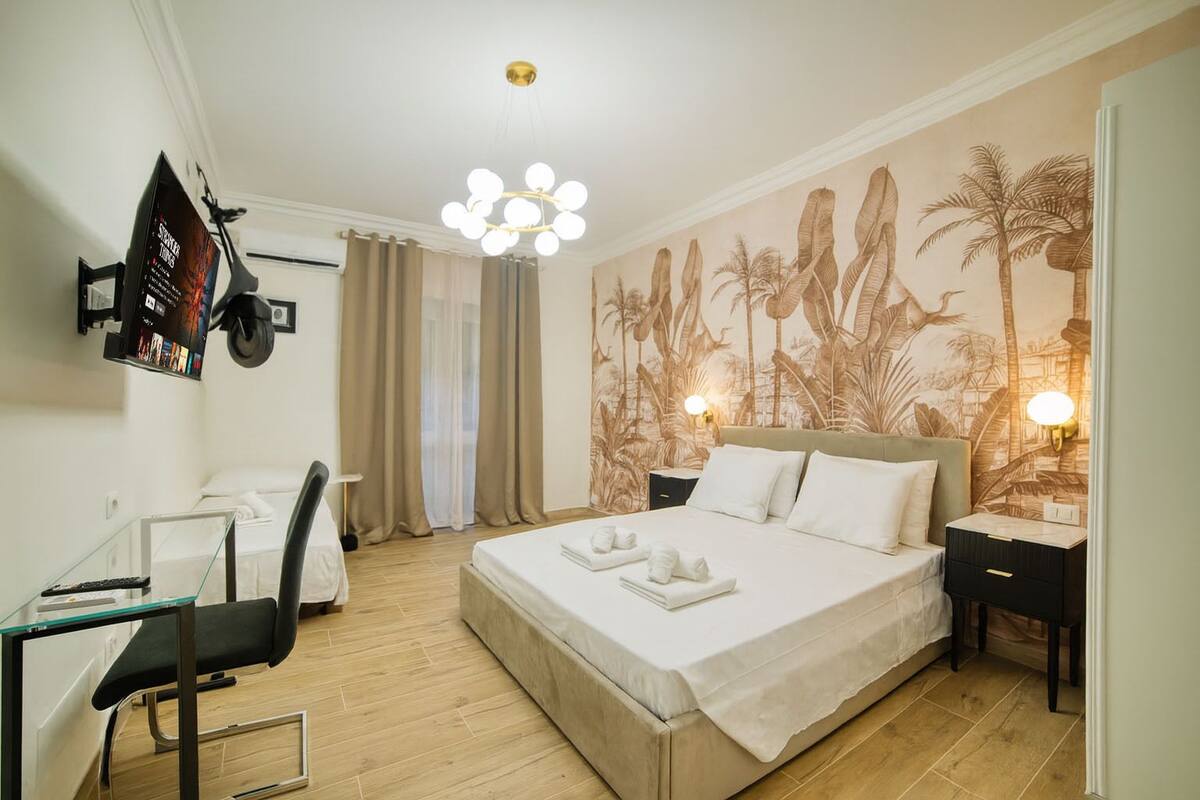 A hotel room with a bed, a desk with a chair, a TV mounted on the wall, and a tropical-themed wall mural.