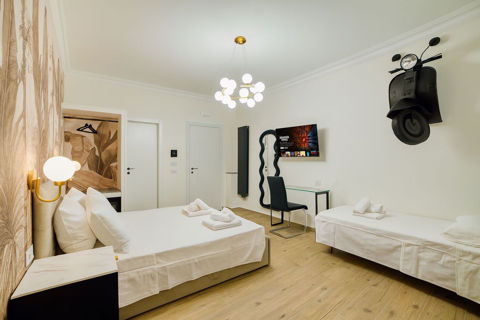 A modern hotel room with two single beds, a desk, a chair, a TV, and a scooter.