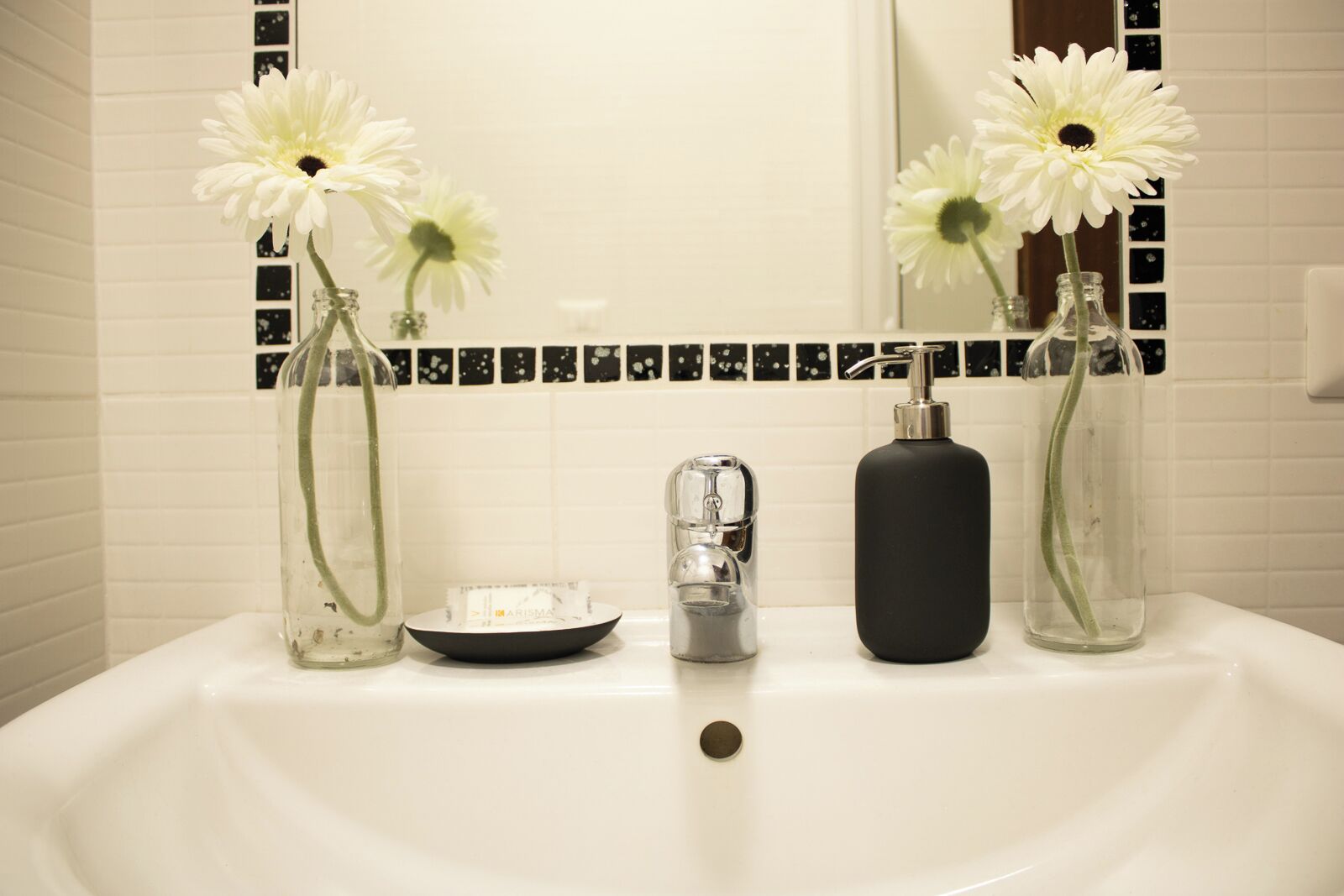 Un lavabo de salle de bain avec trois vases en verre contenant des fleurs blanches, un distributeur de savon et un petit récipient.