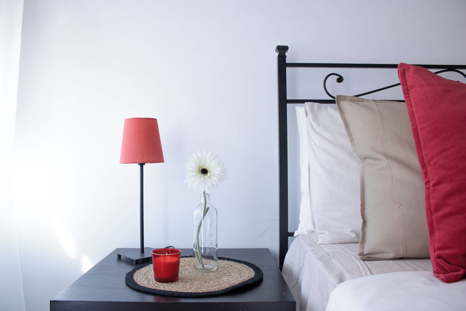 Un dormitorio ordenado con una cama, una mesita de noche, una lámpara, un florero con una flor y una vela roja.