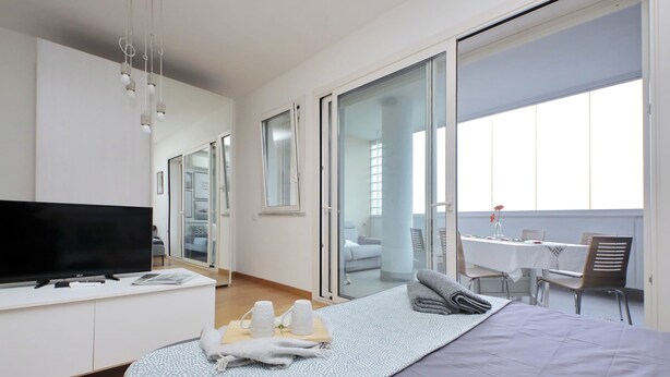 A modern hotel room with a large bed, a flat-screen TV, a desk with a chair, and a balcony with a table and chairs. - Annasole's flat by Domus&Dimore (Rome)