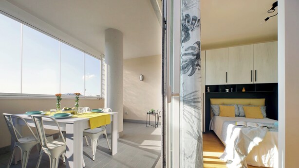 A modern interior with a dining area featuring a white table and chairs, a bed with a yellow headboard, and a view through a glass door.