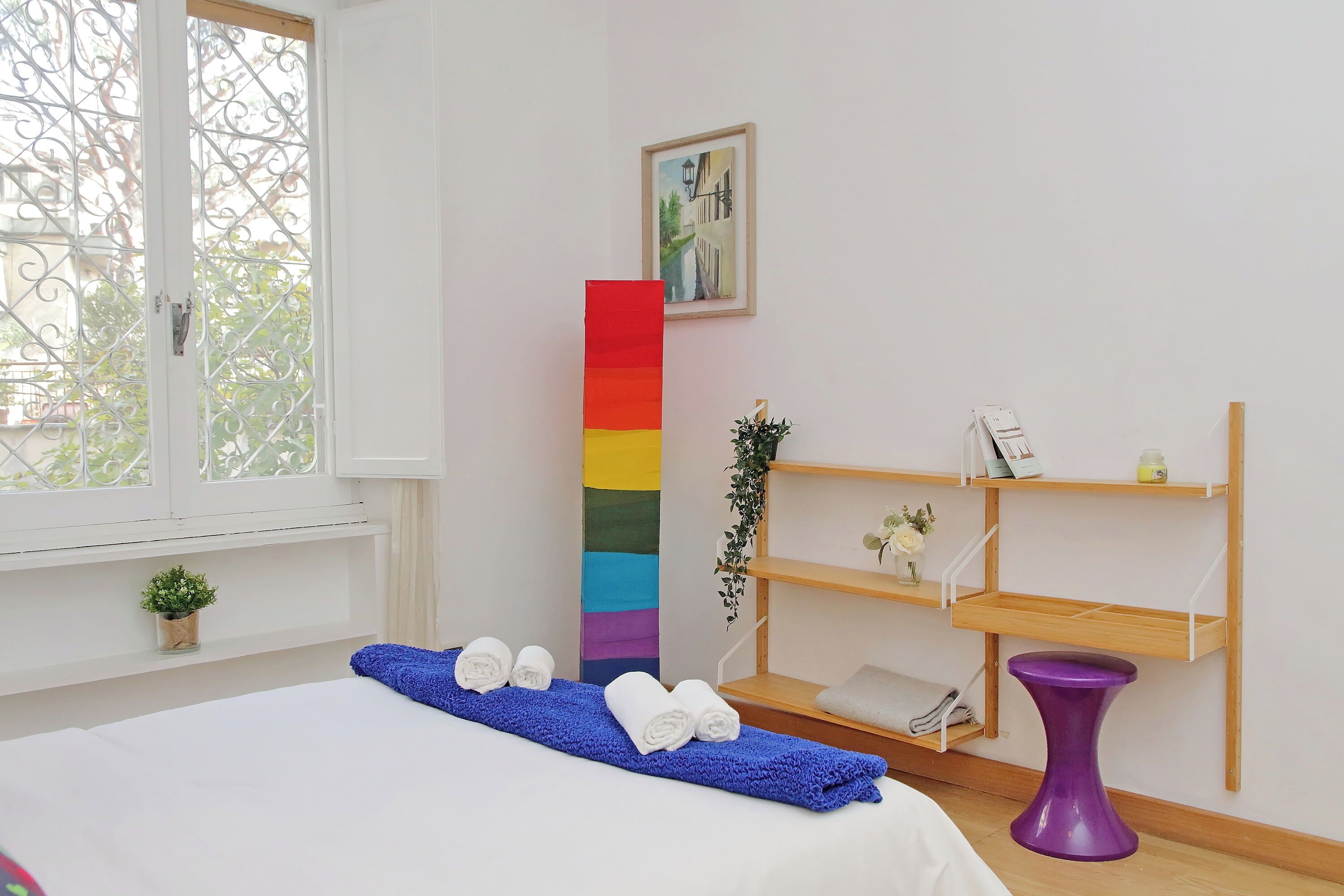 A hotel room with a bed, a blue towel, a tall striped column, wooden shelves, and a purple stool.
