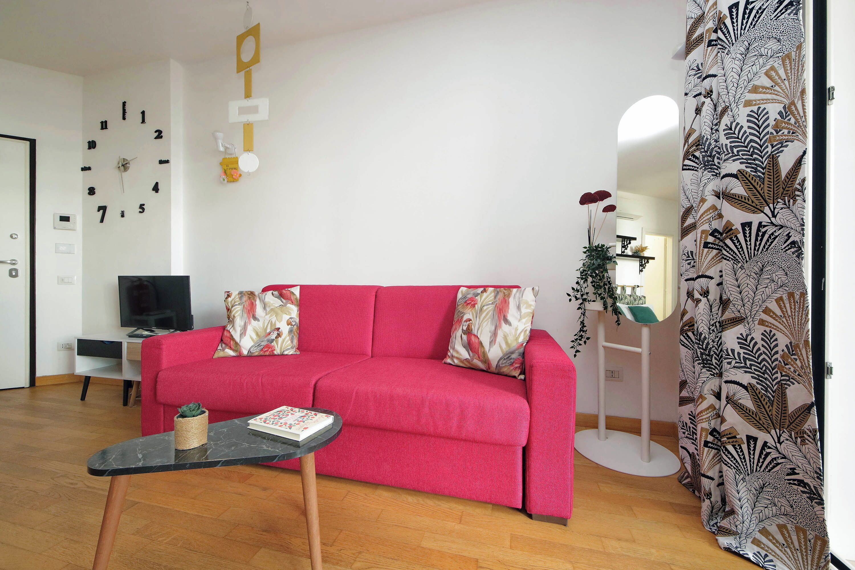 Ein Wohnzimmer mit einem roten Sofa, einem kleinen Tisch mit einem Buch, einem Fernseher und einer Wanduhr.