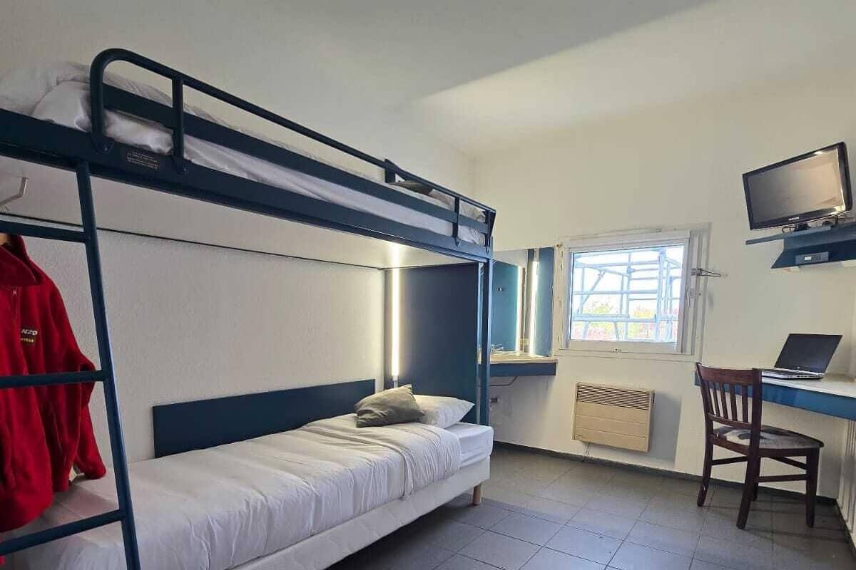 A dormitory room with bunk beds, a desk with a computer, a chair, and a TV.