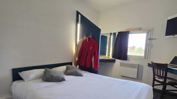 A hotel room with a large bed, a desk with a chair, a mirror, and a window with curtains. - Enzo Hotels 1er Prix Dreux (Dreux)