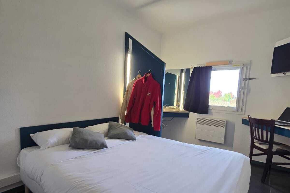 A hotel room with a large bed, a desk with a chair, a mirror, and a window with curtains.