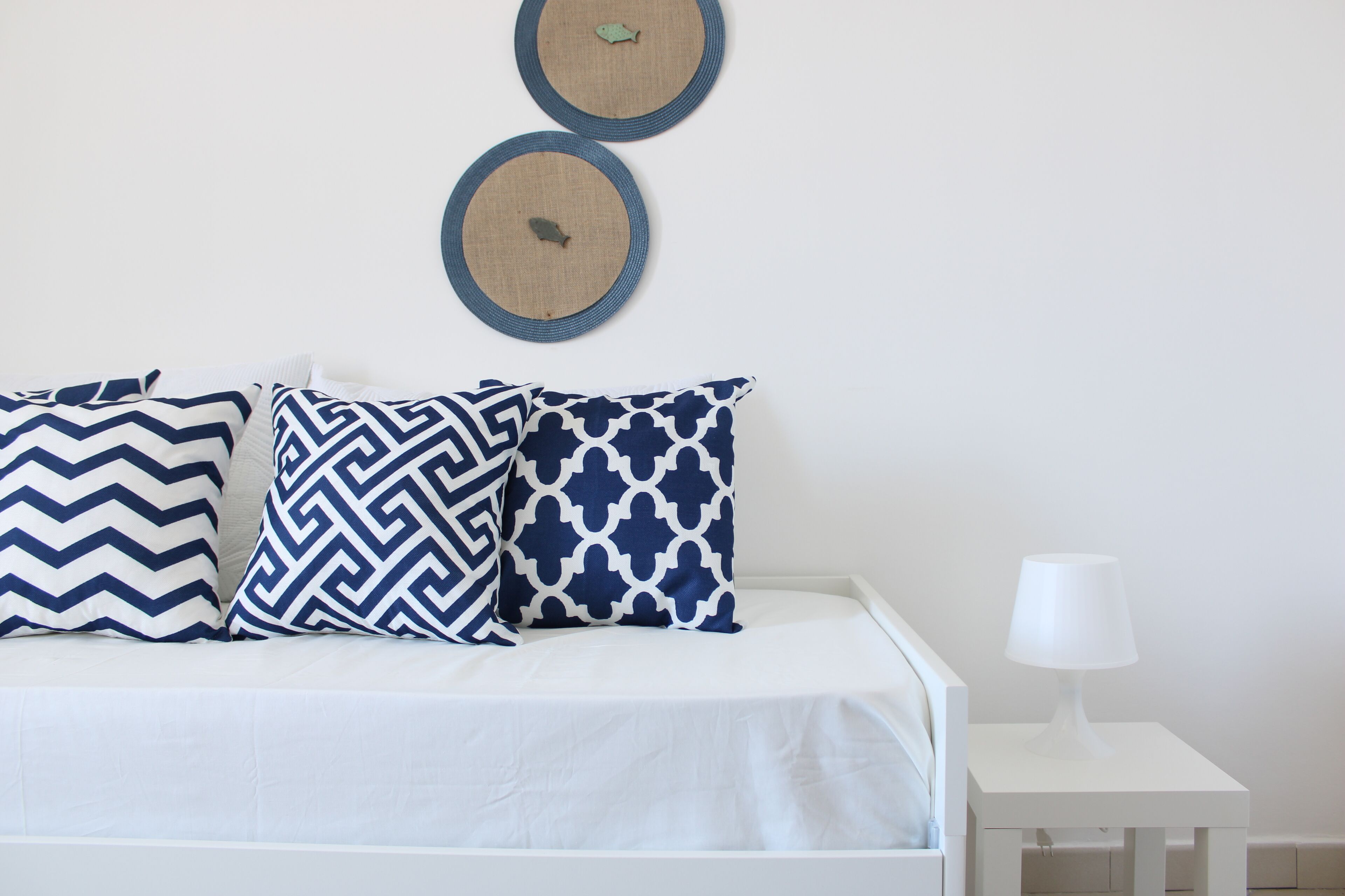 A bed with white bedding and three patterned pillows, a small white nightstand with a lamp, and two wall decorations above the bed.