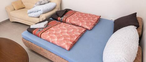 A bed with a blue mattress, two pillows, a blanket, and a chair with cushions.