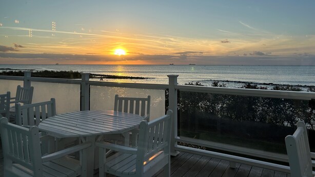 A deck with a table and chairs overlooking a sunset over the ocean.