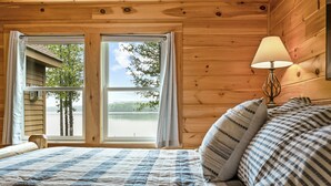 Un dormitorio de cabaña de madera con una cama grande, una mesita de noche con lámpara y una ventana con cortinas.