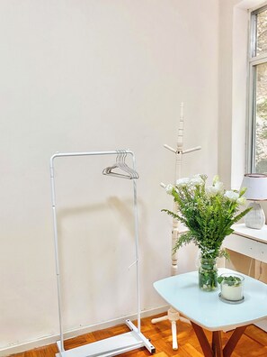 Een kamer met een metalen kledingrek, een klein tafeltje met een vaas en een kaars, en een raam met uitzicht.