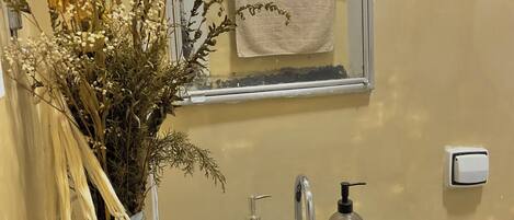 Un baño con lavamanos, espejo, toallero y un florero con flores secas.