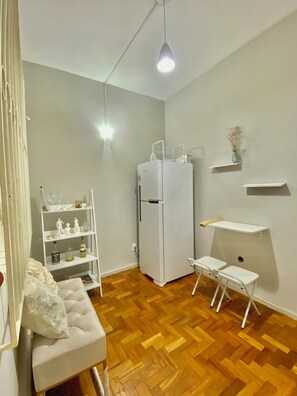 Een knus woongedeelte met een witte koelkast, een kleine witte tafel met stoelen, een houten visgraatvloer en een hanglamp.