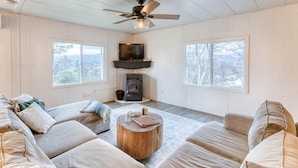A cozy living room with a fireplace, large windows, and a ceiling fan. - Yosemite Boho Bungalow, Pet Friendly + Mtn Views! (Coarsegold)