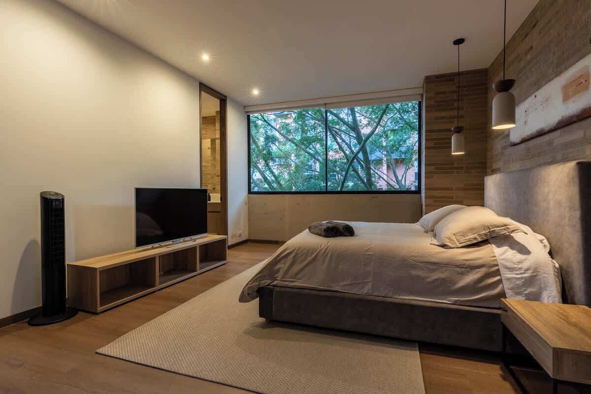 A modern bedroom with a large bed, a TV, and a window with a view of trees.