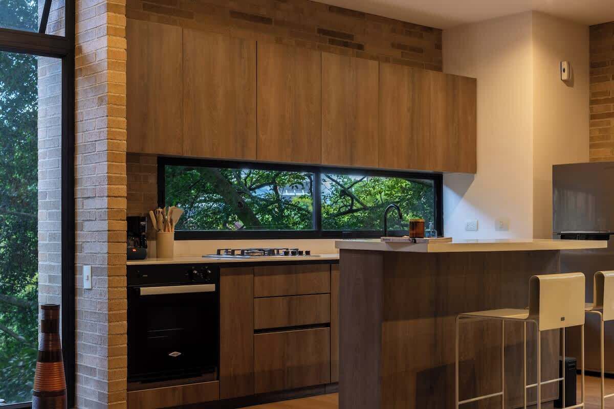 Una cocina moderna con armarios de madera, horno empotrado, ventana con vistas a árboles y taburetes de bar.