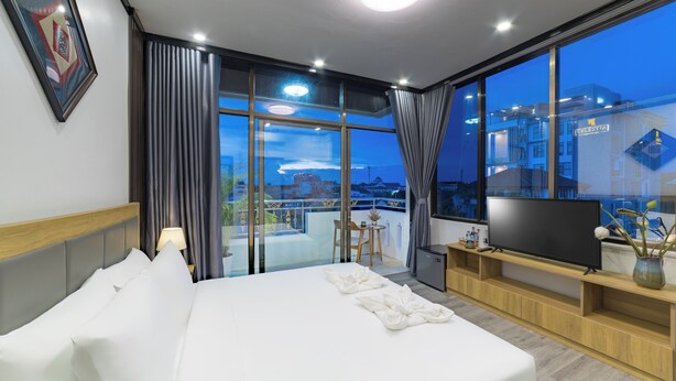 A hotel room with a large bed, a TV, a small table with chairs, and a balcony with a view of the city. - Peaceful 2 Hotel (Vientiane)