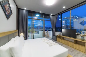 A hotel room with a large bed, a TV, a small table with chairs, and a balcony with a view of the city.