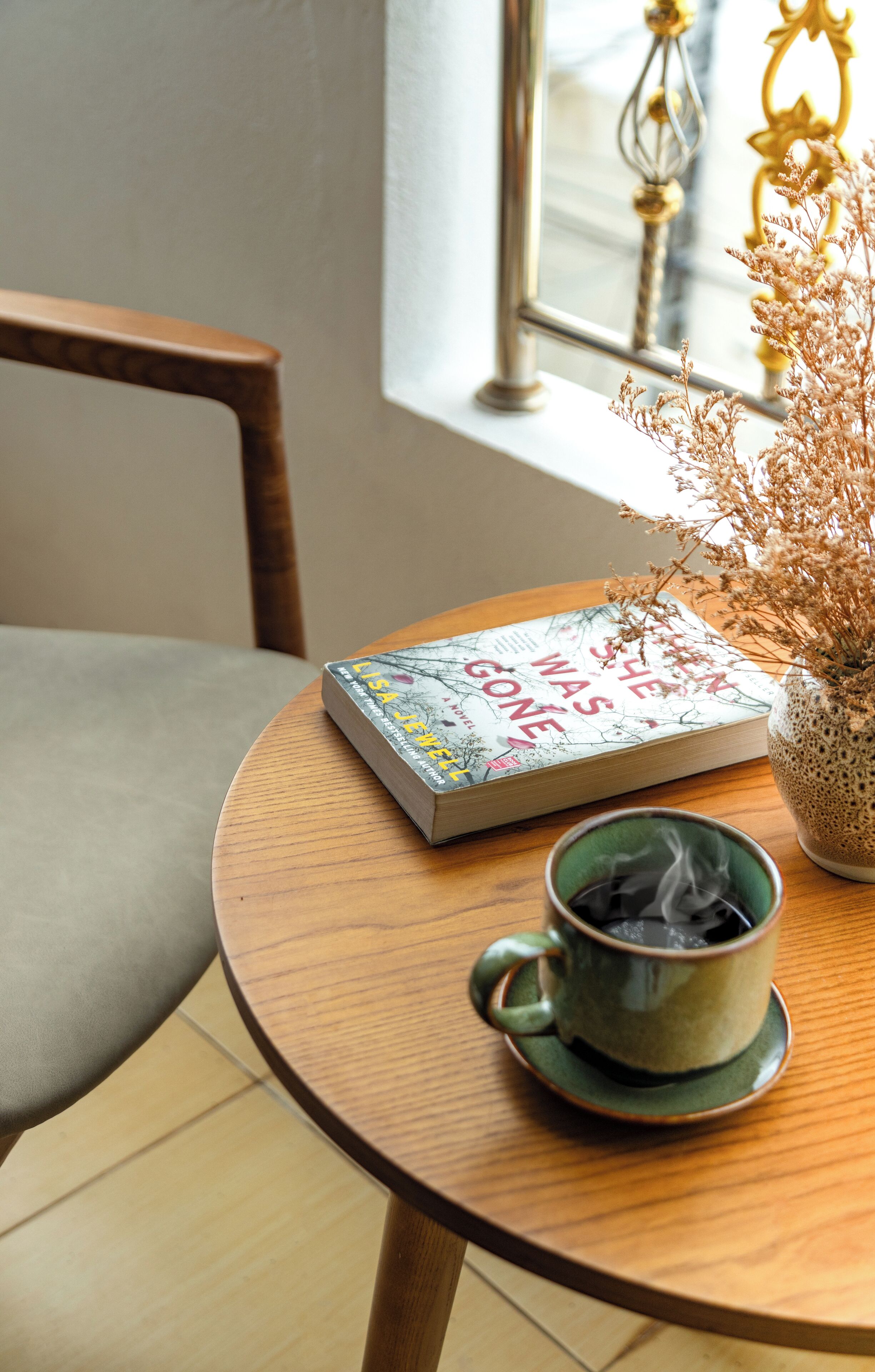 A round wooden table with a book titled "When She Was Gone" and a green ceramic cup with steam.