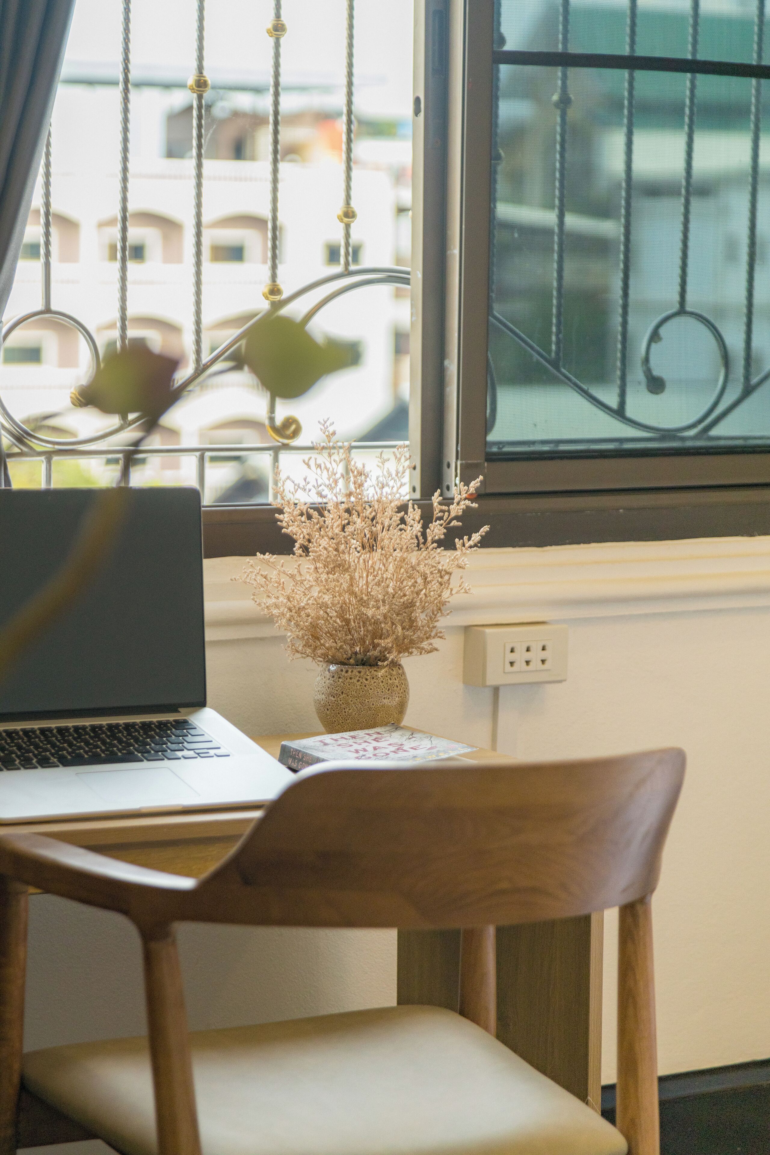 A laptop on a table next to a wooden chair, with a vase of dried flowers on the windowsill.