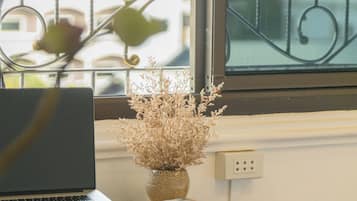 Un portátil sobre una mesa, junto a una silla de madera, con un florero de flores secas en el alféizar de la ventana.