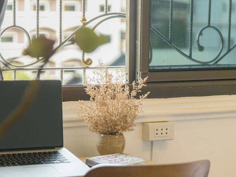 Un portátil sobre una mesa, junto a una silla de madera, con un florero de flores secas en el alféizar de la ventana.