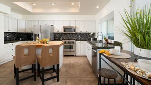 A modern kitchen with white cabinets, a central island with bar stools, a stainless steel refrigerator, and a microwave. - Luxury Palm Desert Condo: Pool & Mountain Views (Palm Desert)